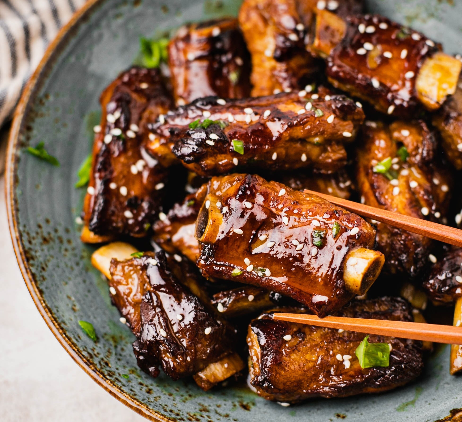 Sweet and sour pork ribs coated in a glossy, caramelized sauce, garnished with sesame seeds and chopped green onions, served in a bowl. Tender, fall-off-the-bone ribs with a sticky, rich glaze that balances tangy and sweet flavors. There is a pair of chopsticks holding a piece of ribs.