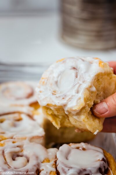 a hand is picking up an eggless cinnamon bun from a full baking dish. The cinnamon rolls are topped with vegan frosting