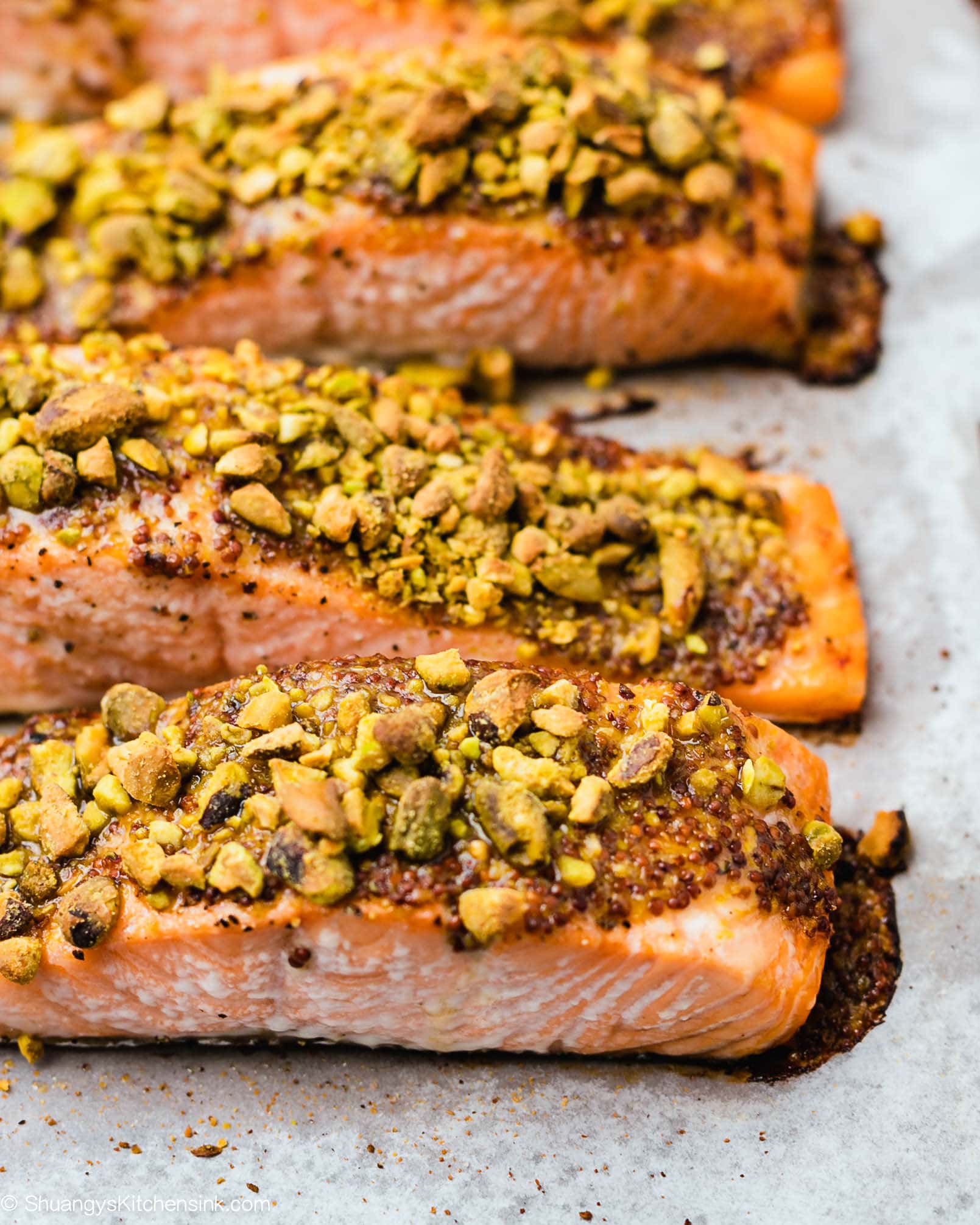 salmon filets on a baking sheet before and after being oven baked. The salmon is topped with crushed pistachios, honey, and mustard