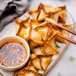 A pair of chopsticks is grabbing a crab rangoon and is going to dip it in a soy dipping sauce