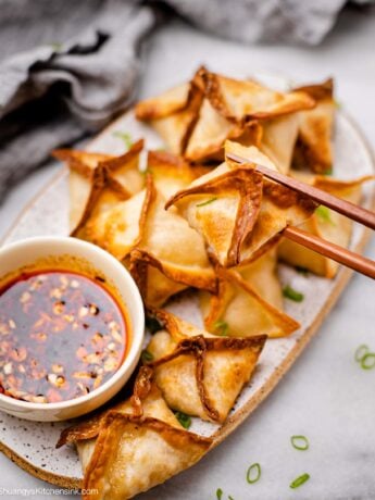 A pair of chopsticks is grabbing a crab rangoon and is going to dip it in a soy dipping sauce