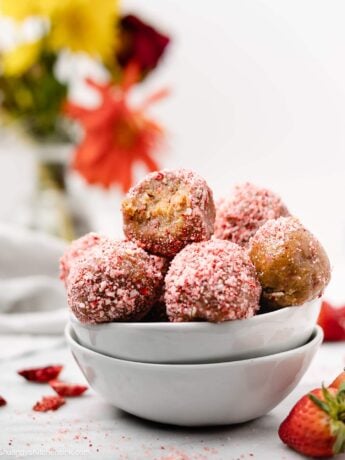 A white small dish with pink no bake strawberry crunch energy balls. they are coated in freeze-dried strawberries and coconut