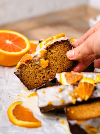 a hand is reaching for a homemade paleo orange loaf slice. it's topped with a vegan icing and fresh citrus