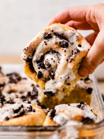 A hand is reaching and grabbing a fluffy baked dessert topped with vegan icing and crushed oreos