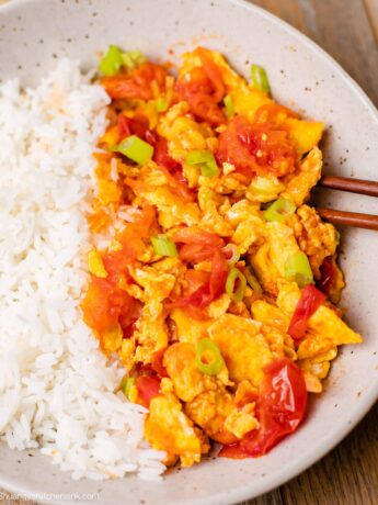 Chinese egg and tomato stir fry serve over rice on a plate,with chopstick resting on the edge of the dish