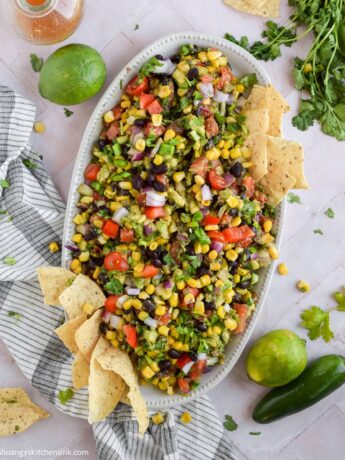 Cowboy Caviar mixed together in a white bow. It is full of bell peppers, onion, beans, corn, avocado, fresh herbs, and a flavorful dressing. There are a few tortilla chips that are sitting on the side of the bowl.