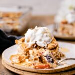 A slice of peach lasagna is on a plate and topped with nuts and a whipped topping. A fork has cut a bite out of it.