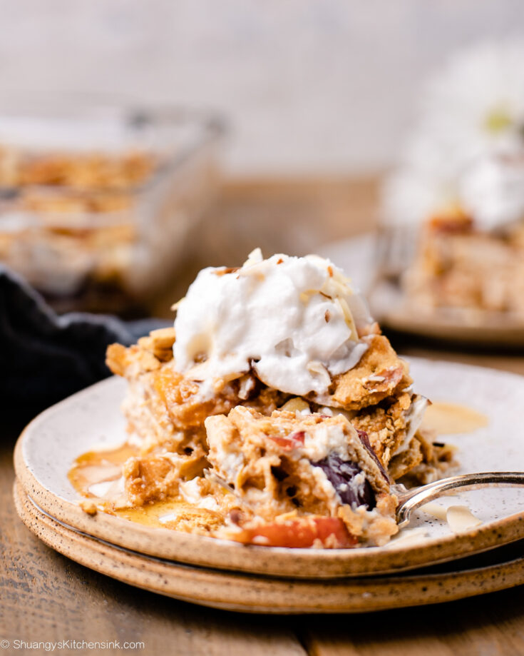 A slice of peach lasagna is on a plate and topped with nuts and a whipped topping. A fork has cut a bite out of it.