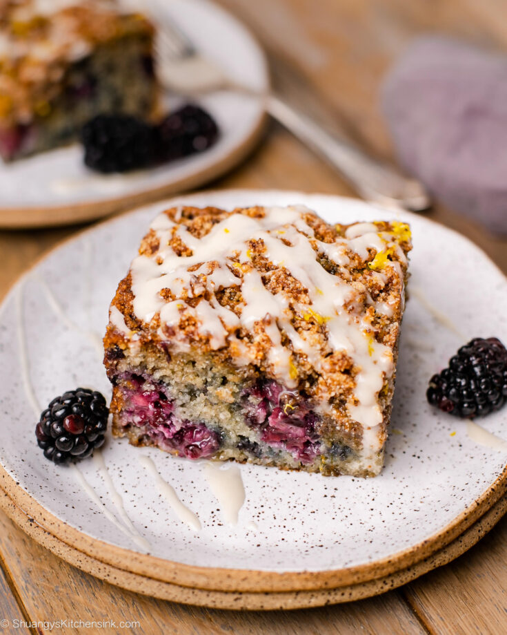 A square of the coffee cake has blackberry swirl and glaze on top, while sitting on a stack of small plates