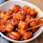 This image shows a bowl full of crispy gochujang tofu with sesame seeds on top, and a pair of chopsticks reaching for a piece