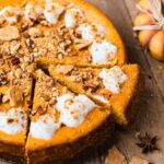 Pumpkin cheesecake is being shown from above, topped with whipped cream, graham, and pecans, with 2 slices cut