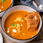 There is a bowl of creamy vegan tomato soup with fresh basil, croutons, and crusty bread