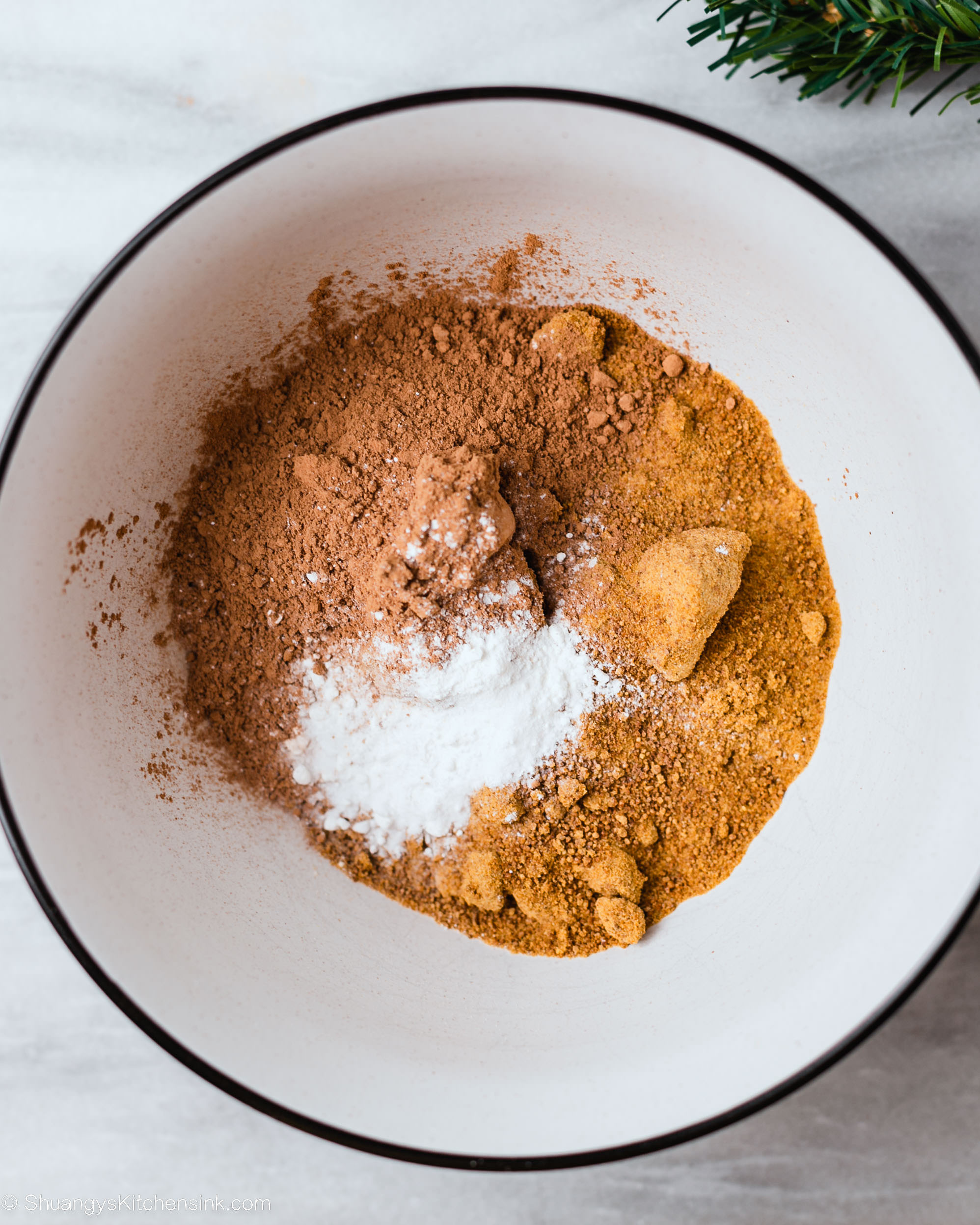 A white bowl with cocoa powder, brown sugar, and baking powder on a white surface—perfect ingredients for a gluten free chocolate Swiss roll—while a small green pine branch peeks from the corner.