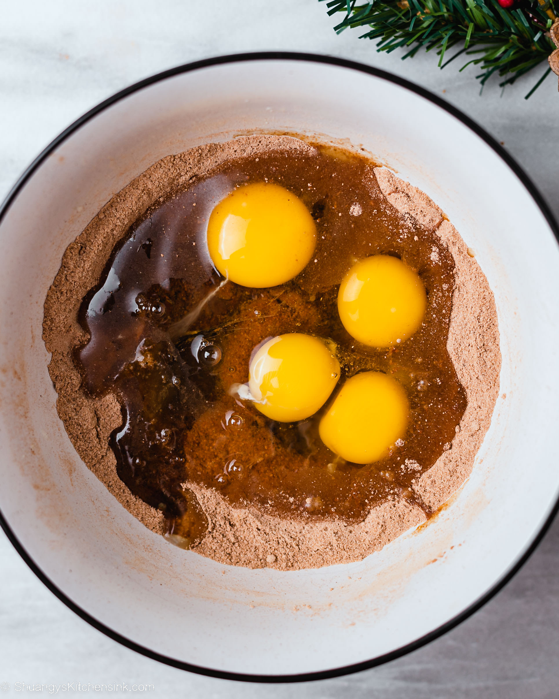 A white bowl holds cocoa powder, four raw eggs, and liquid ingredients for a gluten free chocolate swiss roll on a marble surface, with a sprig of pine partially visible in the corner.