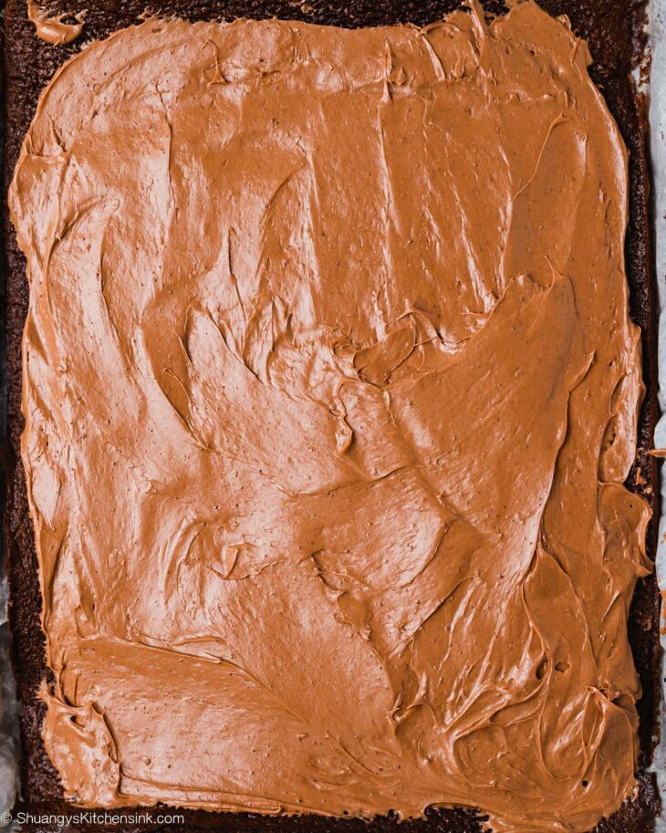 A rectangular sheet of gluten free chocolate cake is covered evenly with creamy chocolate frosting, shown from above. The frosting has smooth, textured swirls spread across the surface.