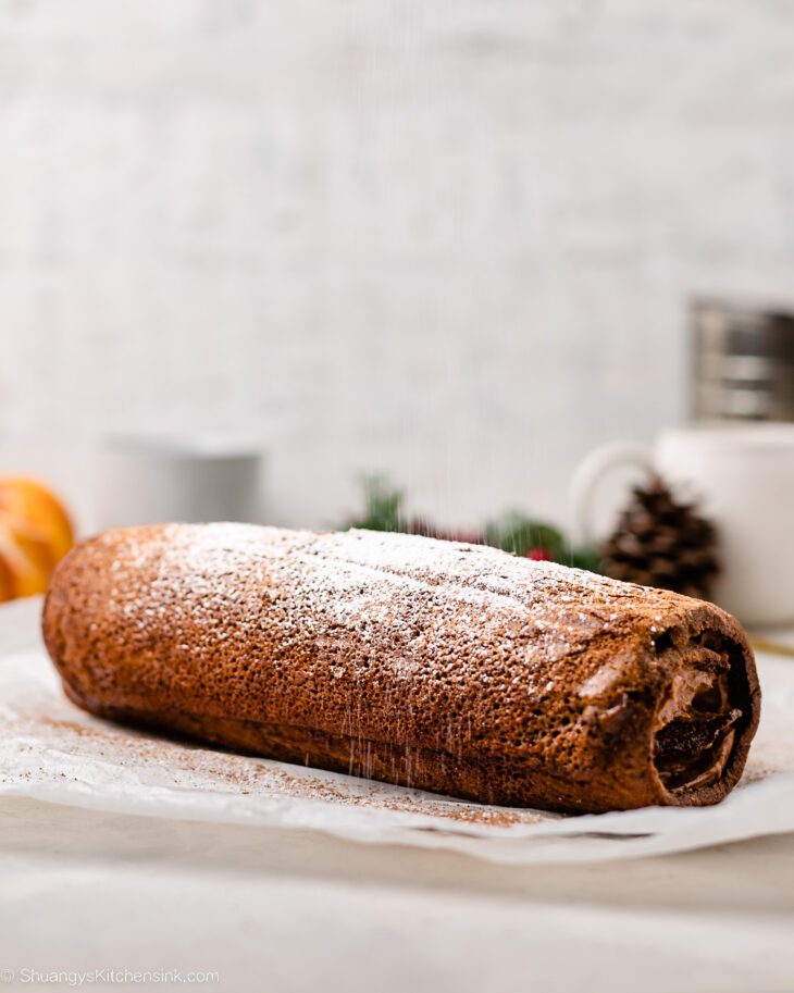 A close-up of a gluten free chocolate swiss roll dusted with powdered sugar, resting on parchment paper with a blurred background featuring a mug, orange slice, pinecone, and festive greenery.
