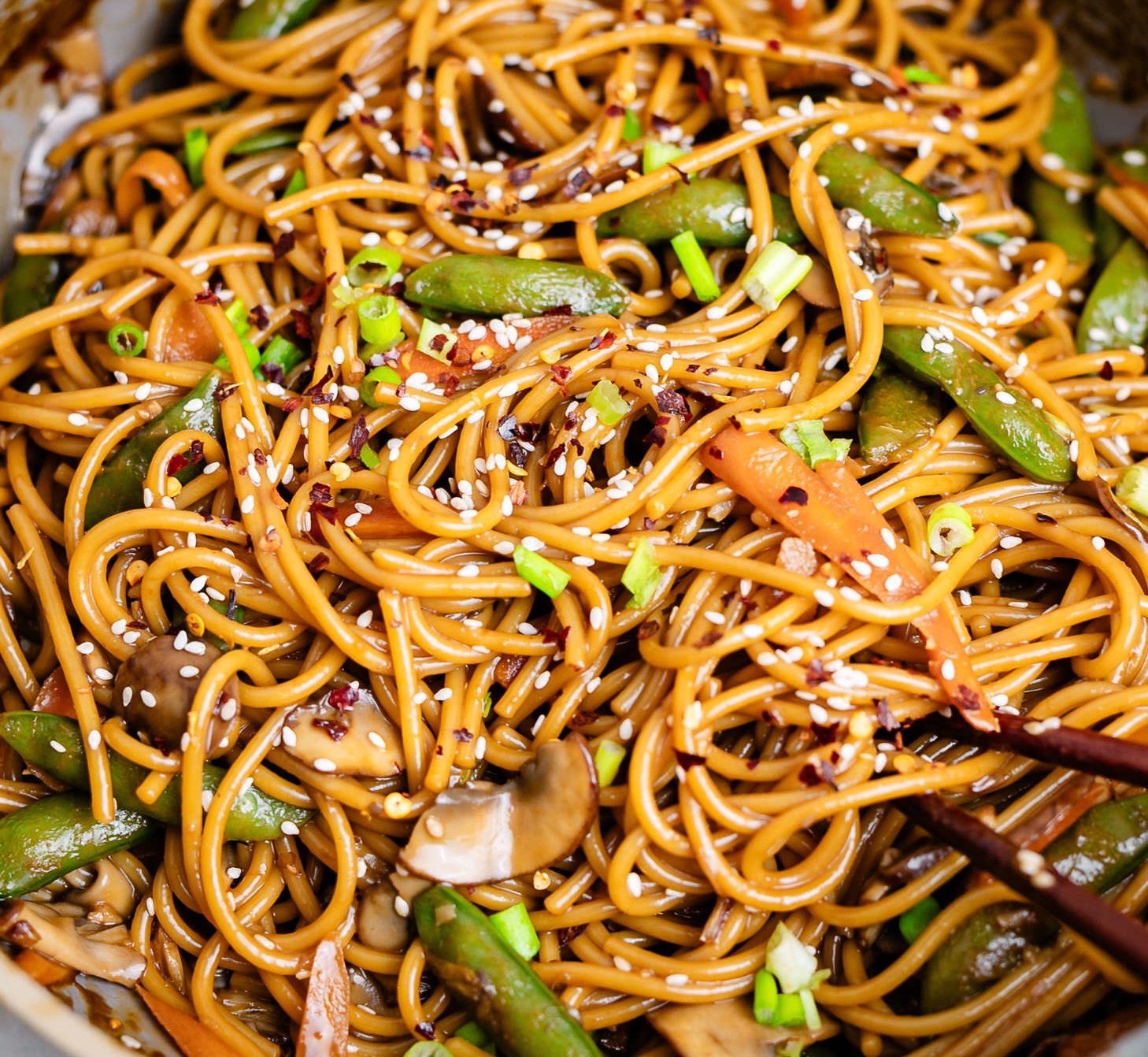 A plate of lo mein with mushroom,snap pees and it is topped with sesame seeds and scallions.