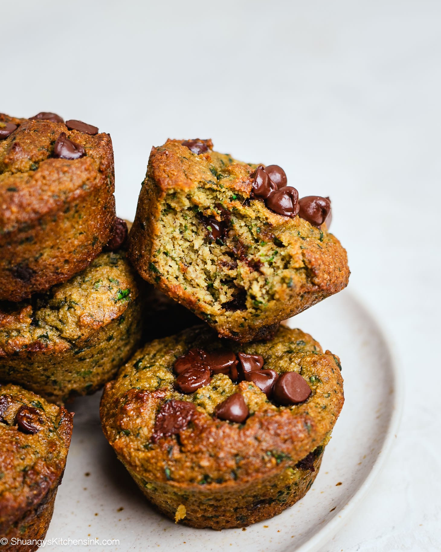 Close-up of almond flour spinach muffins with chocolate chips, showing a moist, tender crumb and golden-brown tops, stacked on a plate with one muffin cut in half.