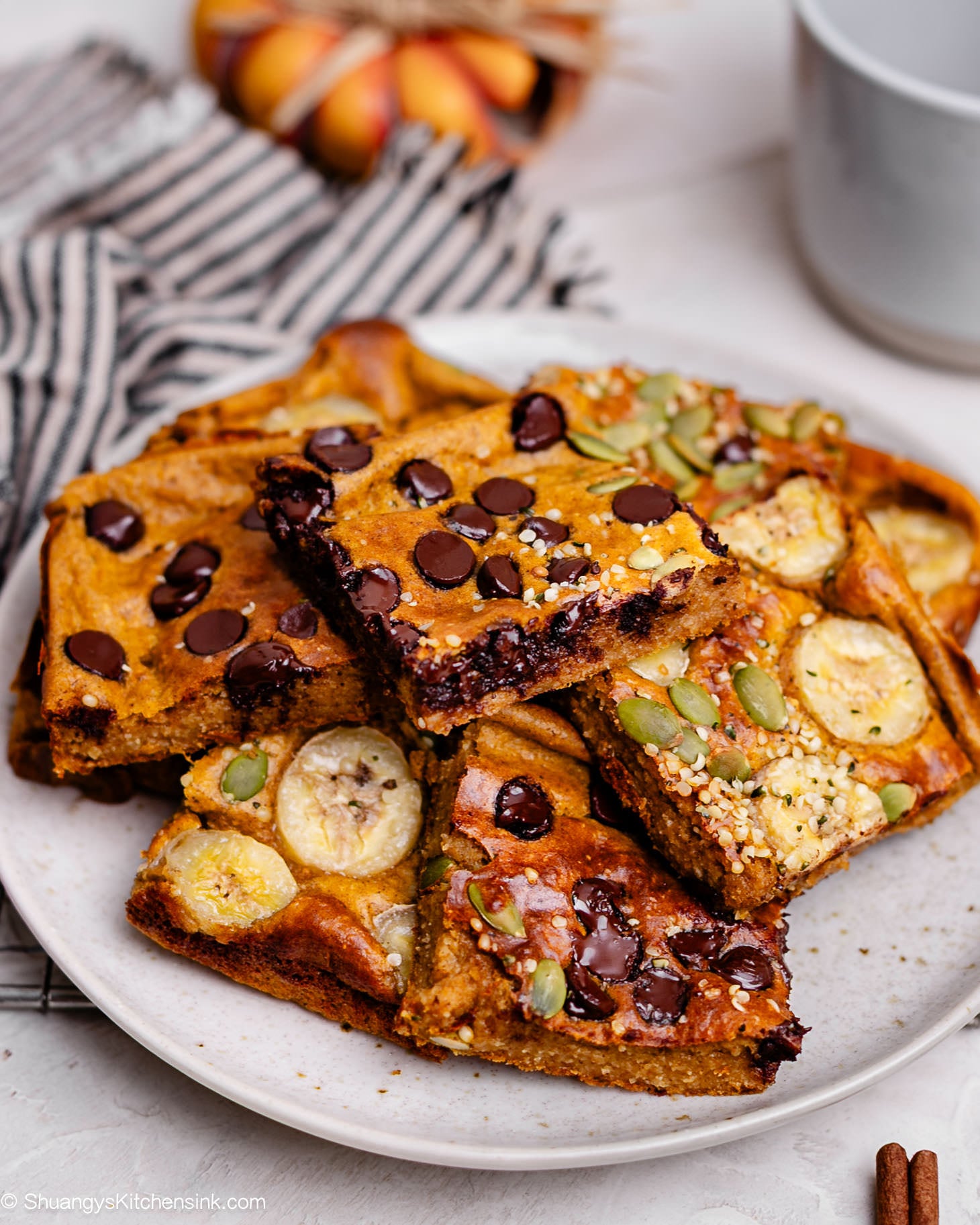 Stack of fluffy pumpkin oat pancakes baked with Greek yogurt, topped with banana slices, chocolate chips, and pumpkin seeds, drizzled with maple syrup on a white plate — cozy fall breakfast idea for busy mornings.