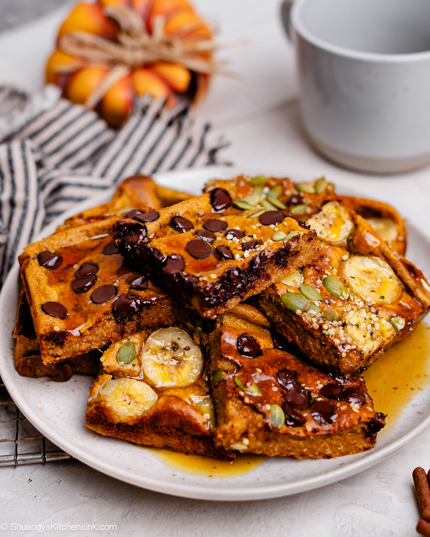 Stack of fluffy pumpkin oat pancakes baked with Greek yogurt, topped with banana slices, chocolate chips, and pumpkin seeds, drizzled with maple syrup on a white plate — cozy fall breakfast idea for busy mornings.