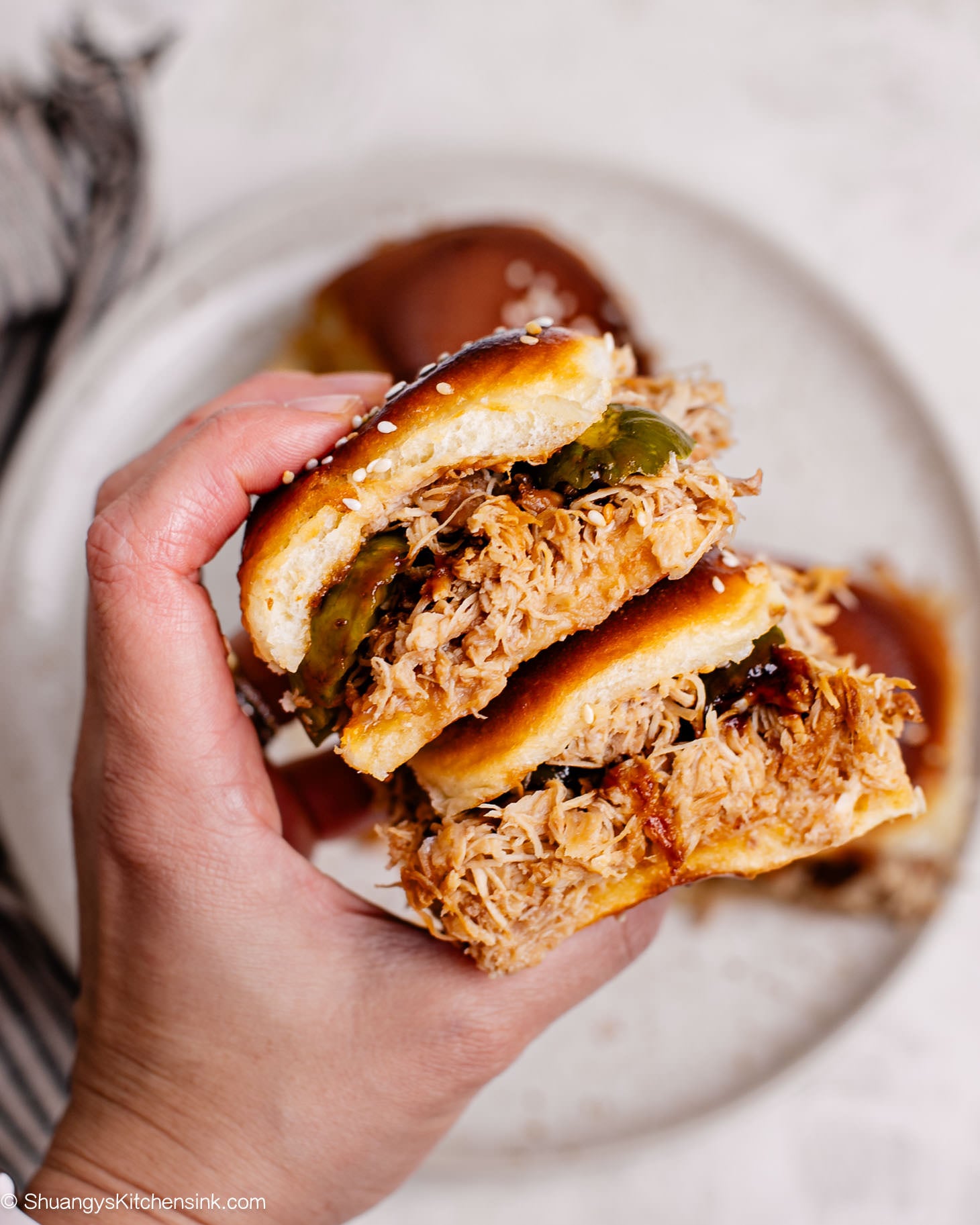 Hand holding a pulled chicken slider made with juicy shredded Asian-style chicken on a toasted sesame bun, with more sliders on a plate in the background.