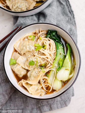 A warm bowl of Chinese wonton soup with frozen wontons, bok choy, and noodles in a savory bone broth, topped with sesame seeds and green onions.