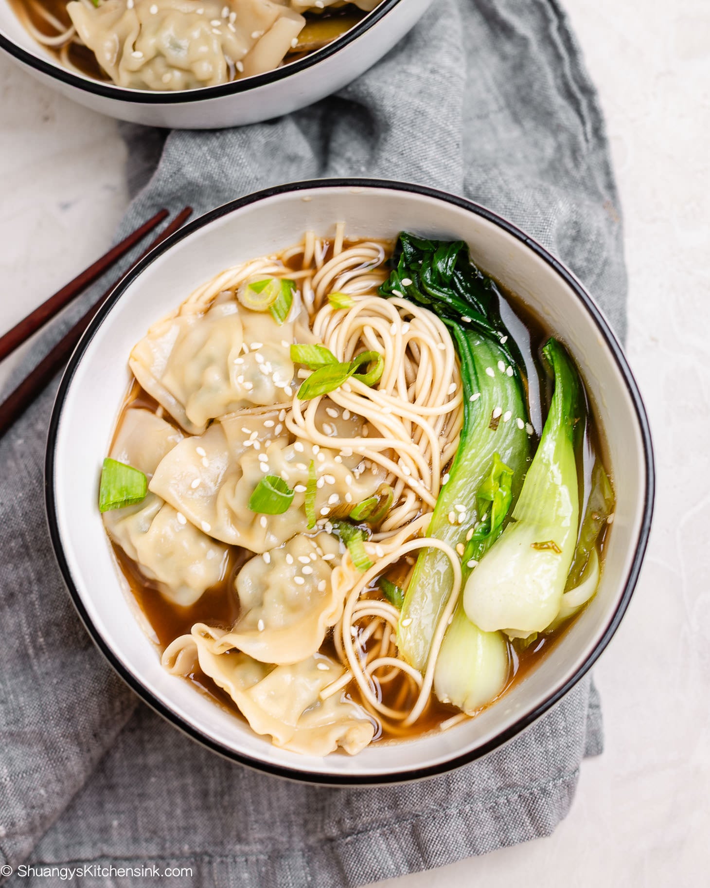 A warm bowl of Chinese wonton soup with frozen wontons, bok choy, and noodles in a savory bone broth, topped with sesame seeds and green onions.
