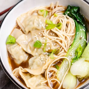 A warm bowl of Chinese wonton soup with frozen wontons, bok choy, and noodles in a savory bone broth, topped with sesame seeds and green onions.