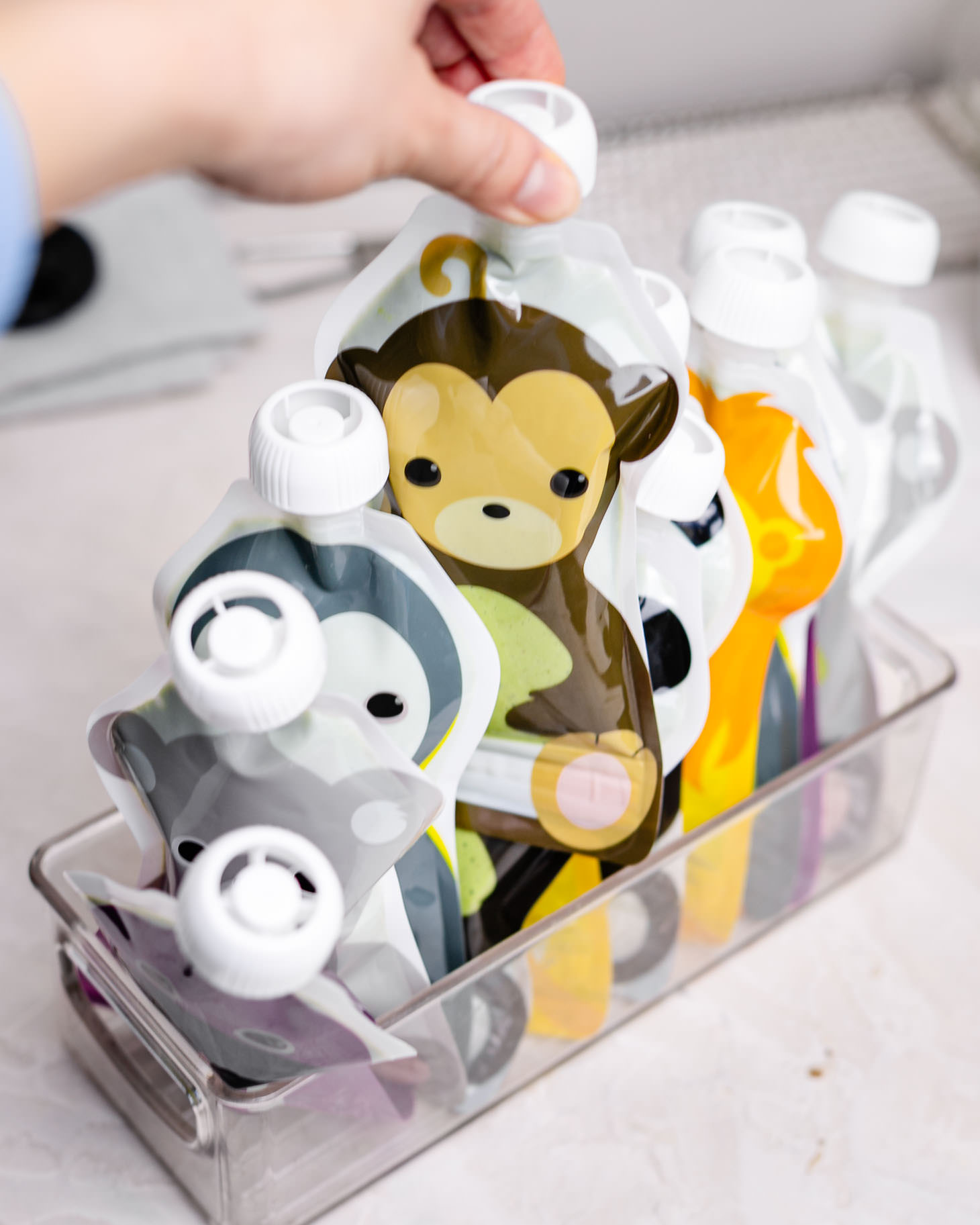 Reusable animal-shaped smoothie pouches filled with homemade kids green smoothie and fruit blends, stored upright in a clear freezer bin for easy grab-and-go snacks.