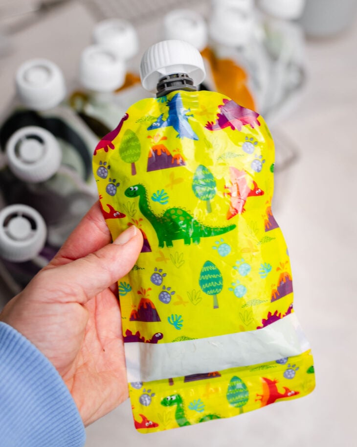 Reusable dinosaur-themed smoothie pouch filled with homemade kids green smoothie, held in hand and ready as a healthy grab-and-go snack.