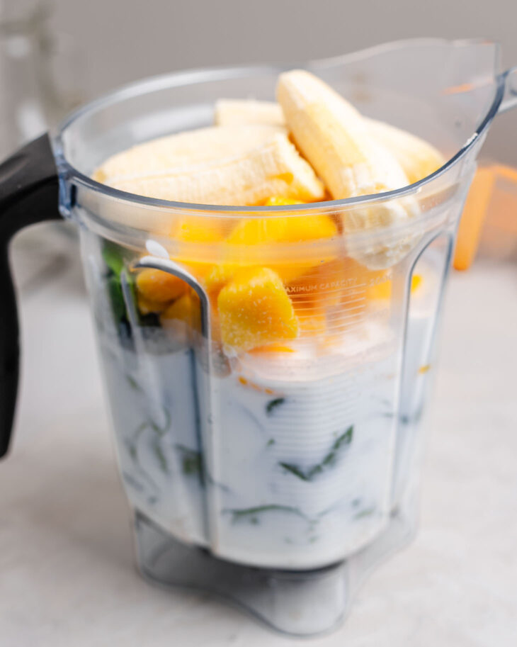 Bananas, frozen mango, spinach, and milk added to a blender pitcher to make a creamy kids green smoothie with spinach.