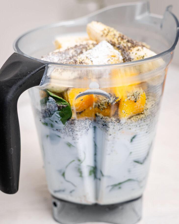Bananas, mango, spinach, milk, chia seeds, and hemp seeds layered in a blender before blending into a nutritious smoothie for kids.