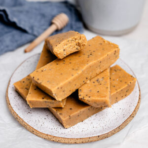 Stack of homemade copycat Perfect Bars on a plate, showing soft, chewy peanut butter almond flour texture with chia seeds, with one bar bitten to reveal the inside, a no-bake healthy protein snack.