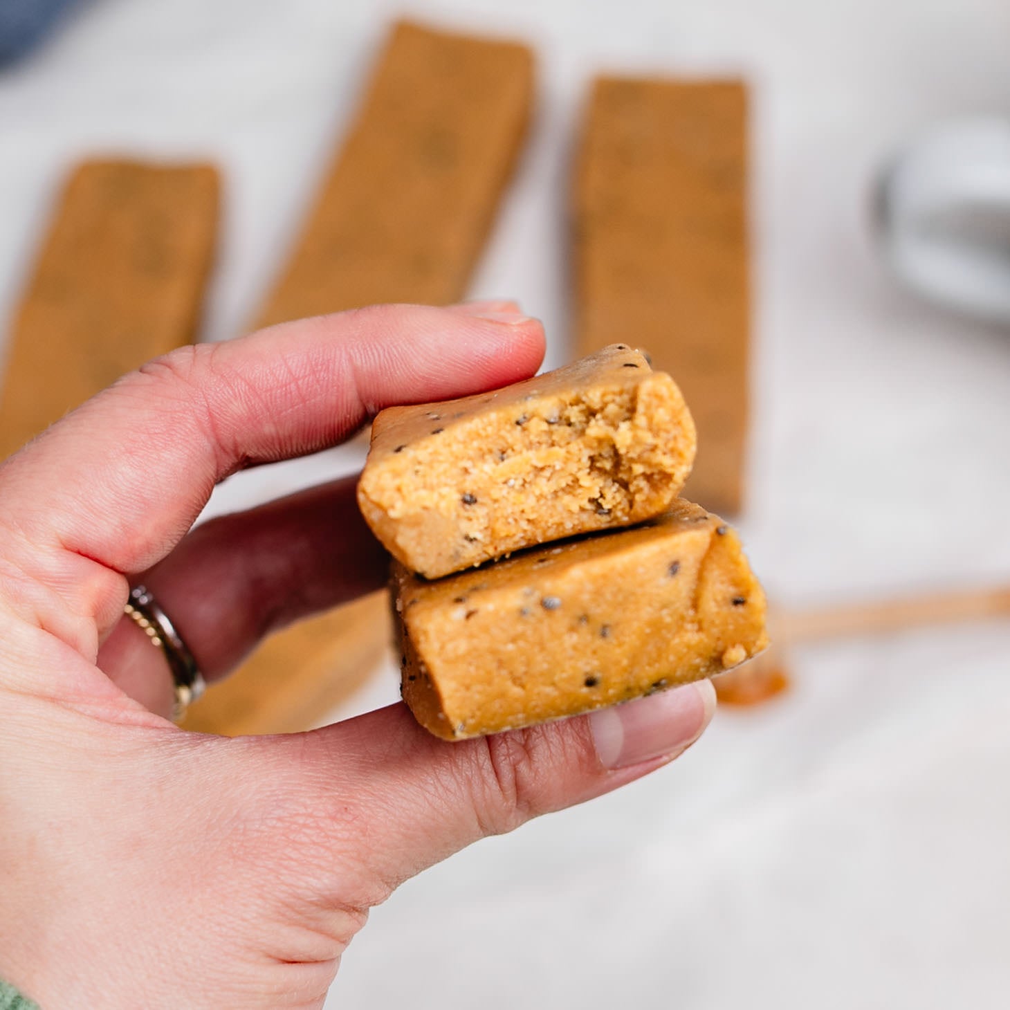 Hand holding two homemade copycat Perfect Bars stacked together, showing a soft, chewy peanut butter almond flour texture with chia seeds inside, with more no-bake protein bars blurred in the background on a white surface.