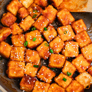 Crispy pan-fried sweet and sour tofu cubes coated in a glossy sauce, garnished with sesame seeds and chopped scallions, cooked in a skillet.