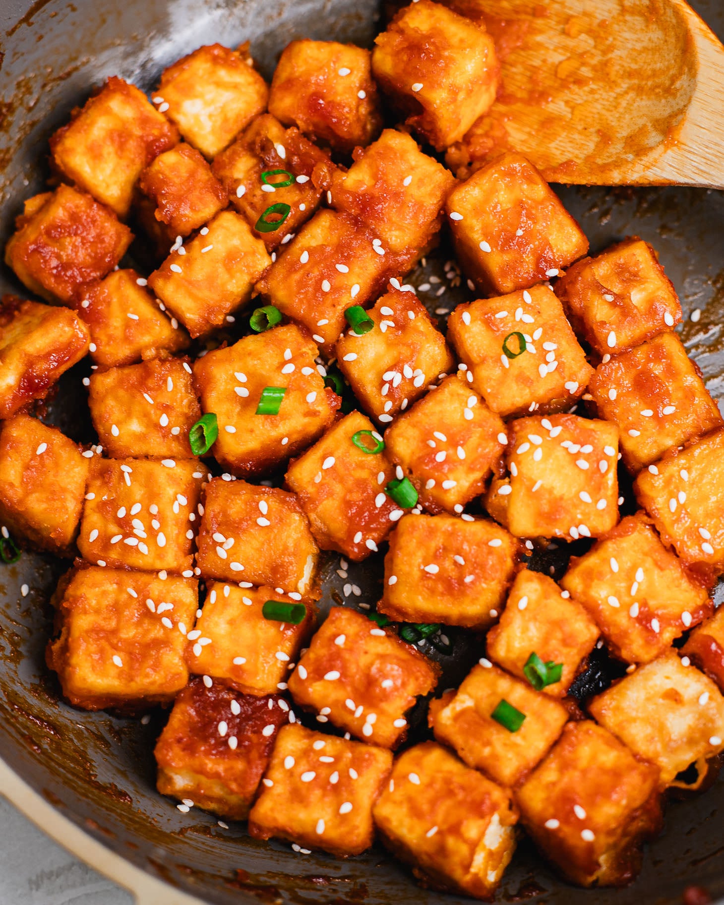 Crispy pan-fried sweet and sour tofu cubes coated in a glossy sauce, garnished with sesame seeds and chopped scallions, cooked in a skillet.