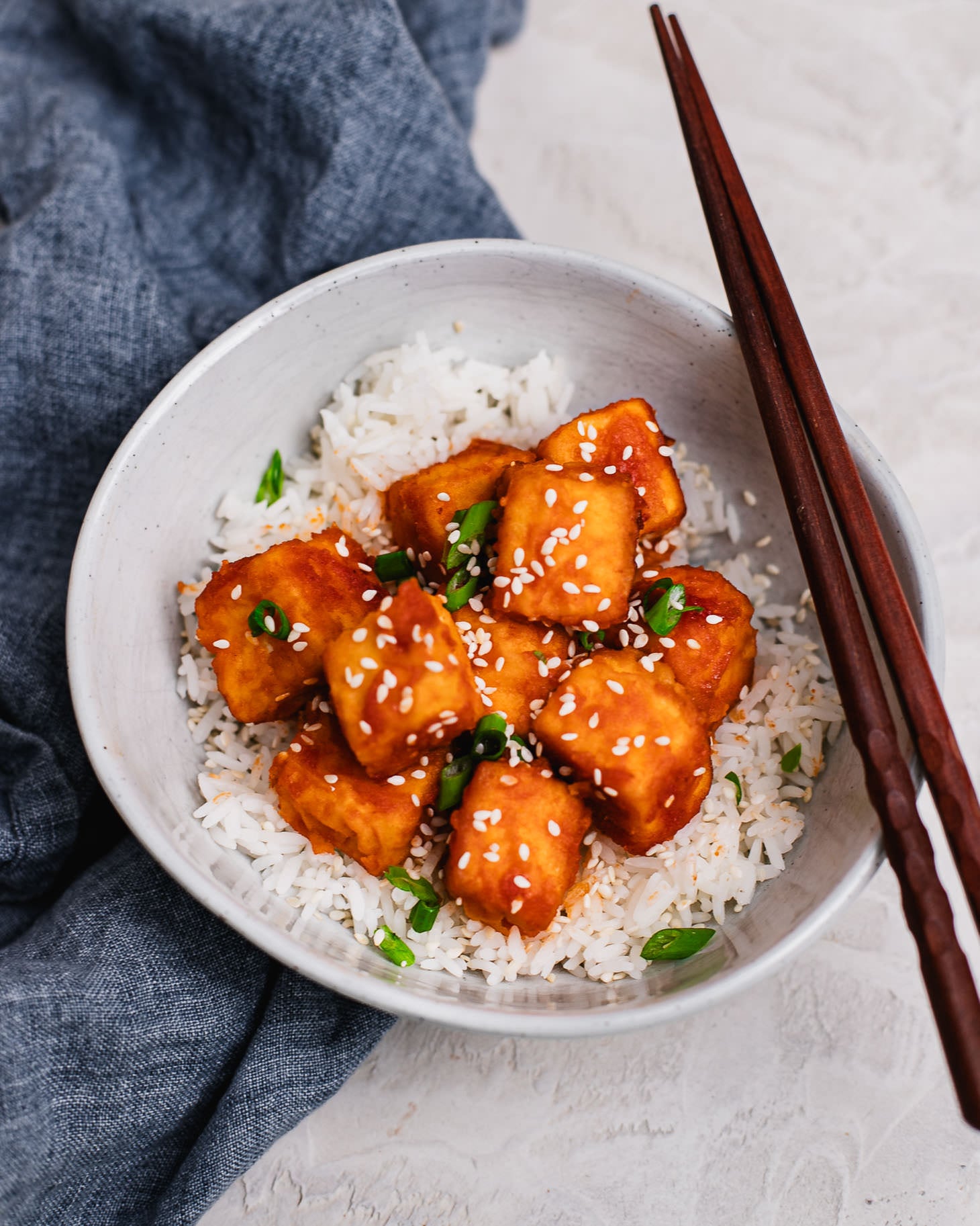 Crispy sweet and sour tofu cubes served over jasmine rice in a bowl, topped with sesame seeds and chopped scallions.