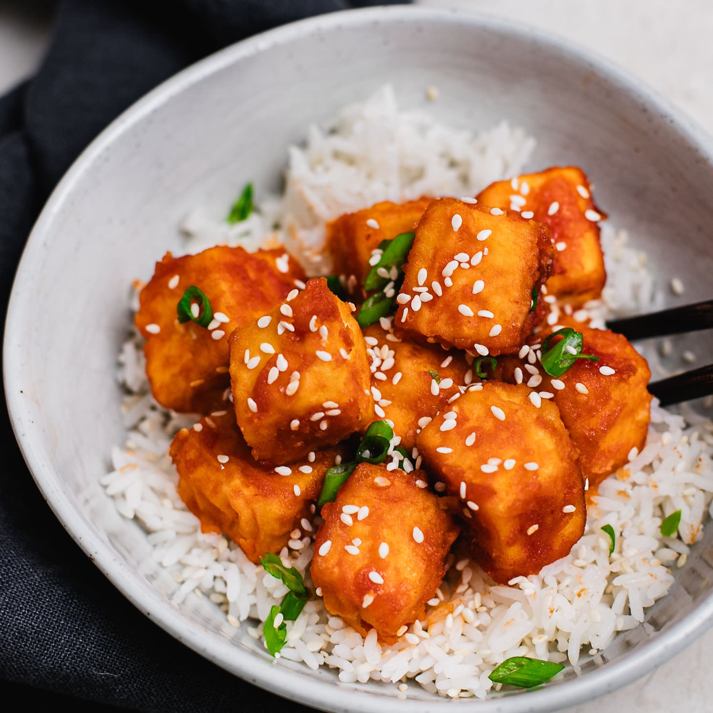 Crispy sweet and sour tofu cubes served over jasmine rice in a bowl, topped with sesame seeds and chopped scallions.