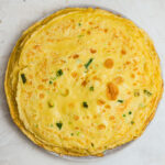 Stack of homemade Chinese Egg Pancakes (鸡蛋饼) on a plate, featuring golden brown spots, tender edges, and fresh scallions throughout. These savory Chinese crepe-style pancakes are soft, chewy, and a traditional comfort breakfast made with eggs, flour, and scallions.
