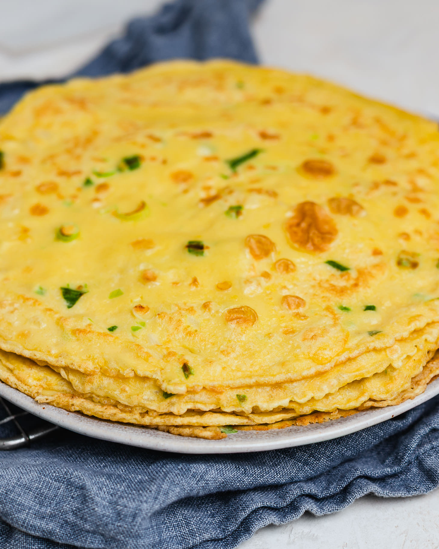 Stack of homemade Chinese Egg Pancakes (鸡蛋饼) on a plate, featuring golden brown spots, tender edges, and fresh scallions throughout. These savory Chinese crepe-style pancakes are soft, chewy, and a traditional comfort breakfast made with eggs, flour, and scallions.