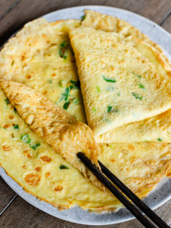Chinese Egg Pancakes (鸡蛋饼) stacked on a plate, showing their thin, tender crepe-like texture with golden brown spots and fresh scallions throughout. One pancake is rolled up with chopsticks to highlight the soft and flexible texture of this traditional Chinese breakfast recipe.