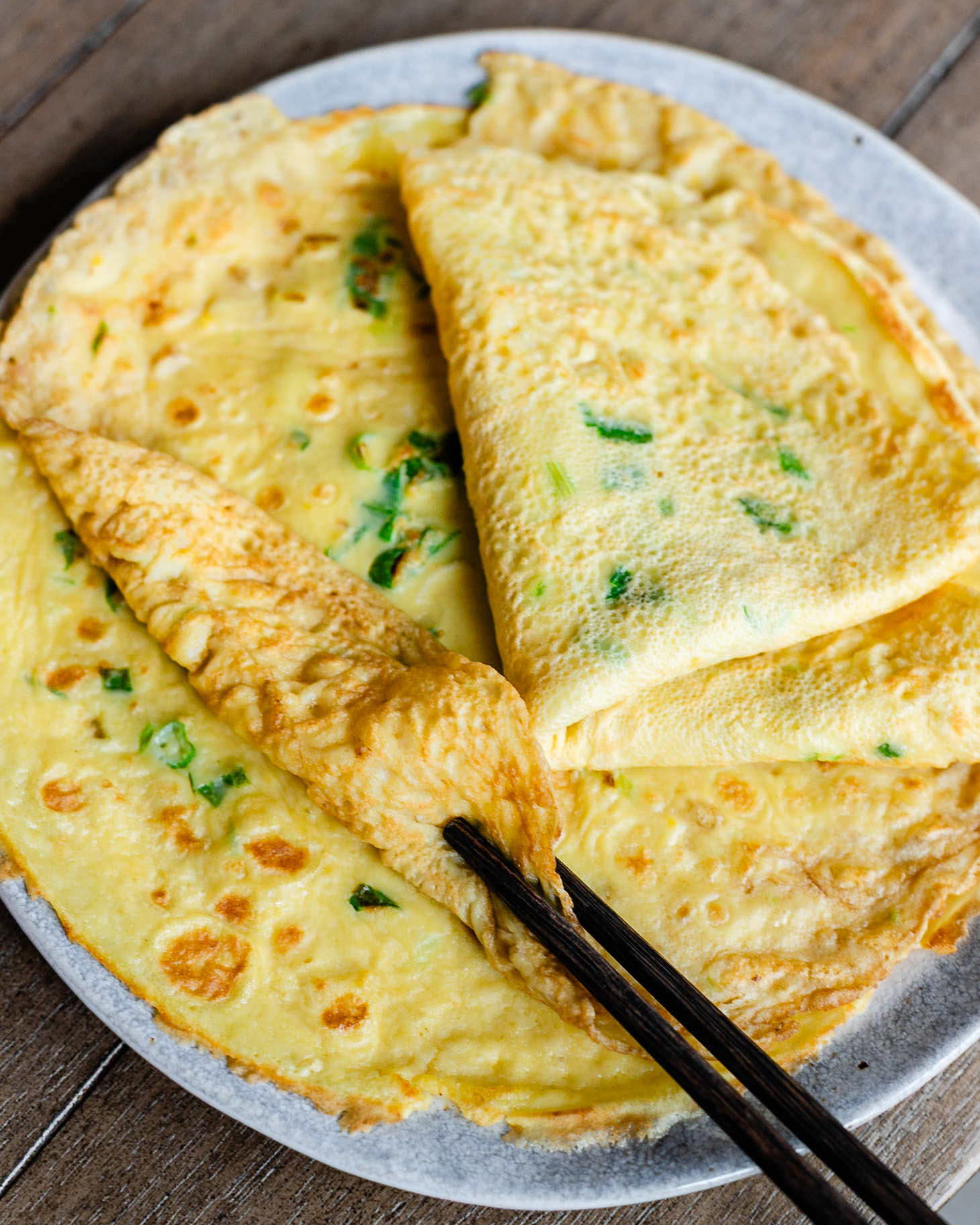 Chinese Egg Pancakes (鸡蛋饼) stacked on a plate, showing their thin, tender crepe-like texture with golden brown spots and fresh scallions throughout. One pancake is rolled up with chopsticks to highlight the soft and flexible texture of this traditional Chinese breakfast recipe.
