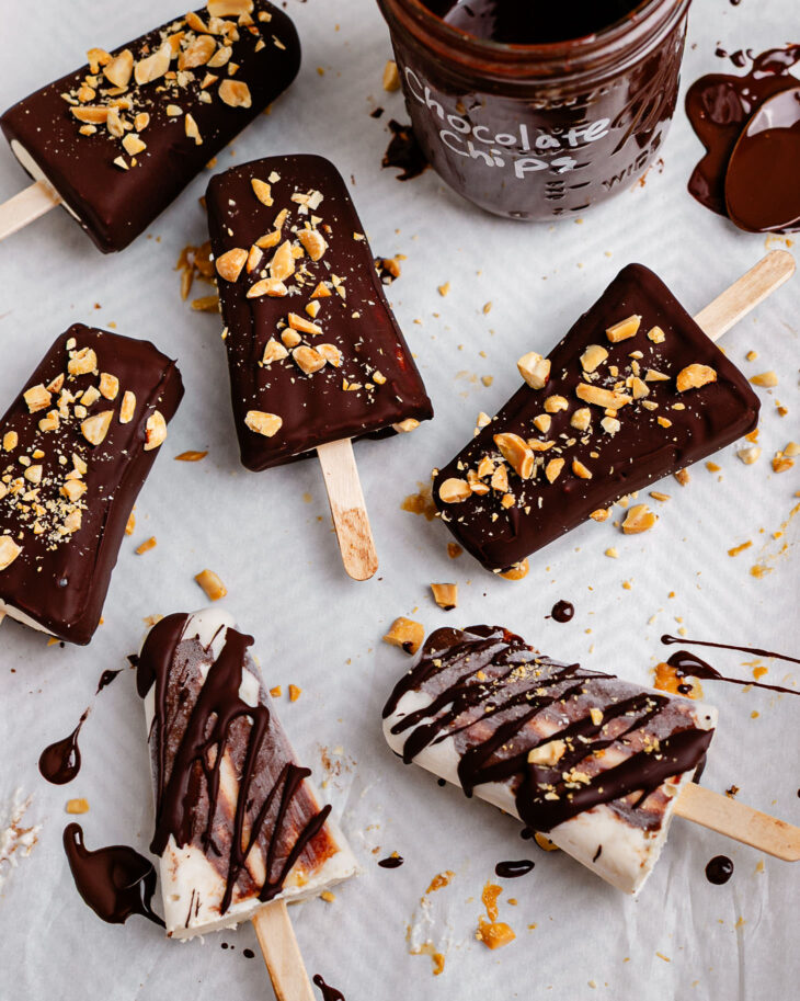 Overhead view of creamy Greek yogurt banana ice cream bars coated in a glossy dark chocolate shell and topped with chopped peanuts on parchment paper. Some bars are fully dipped while others are drizzled with chocolate, with melted chocolate and crushed peanuts scattered around for a homemade frozen dessert look.