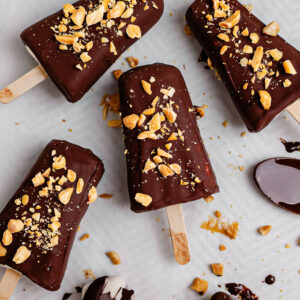 Overhead view of creamy Greek yogurt banana ice cream bars coated in a glossy dark chocolate shell and topped with chopped peanuts on parchment paper. Some bars are fully dipped while others are drizzled with chocolate, with melted chocolate and crushed peanuts scattered around for a homemade frozen dessert look.