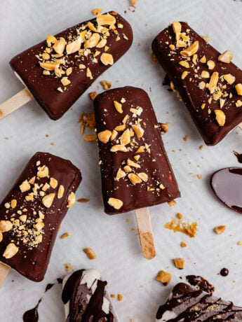 Overhead view of creamy Greek yogurt banana ice cream bars coated in a glossy dark chocolate shell and topped with chopped peanuts on parchment paper. Some bars are fully dipped while others are drizzled with chocolate, with melted chocolate and crushed peanuts scattered around for a homemade frozen dessert look.