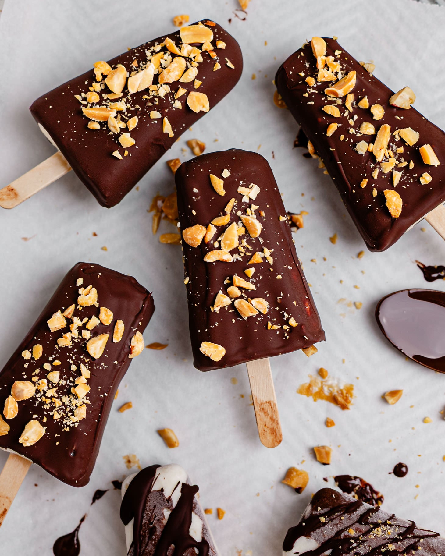 Overhead view of creamy Greek yogurt banana ice cream bars coated in a glossy dark chocolate shell and topped with chopped peanuts on parchment paper. Some bars are fully dipped while others are drizzled with chocolate, with melted chocolate and crushed peanuts scattered around for a homemade frozen dessert look.