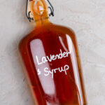 Glass bottle of homemade lavender syrup filled with golden amber syrup and labeled "Lavender Syrup" in white lettering. The bottle sits on a rustic white wooden board scattered with dried lavender buds, alongside a blue linen napkin and coffee mug in the background. A cozy kitchen scene highlighting an easy homemade coffee syrup recipe.