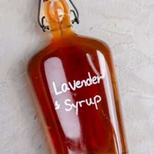 Glass bottle of homemade lavender syrup filled with golden amber syrup and labeled "Lavender Syrup" in white lettering. The bottle sits on a rustic white wooden board scattered with dried lavender buds, alongside a blue linen napkin and coffee mug in the background. A cozy kitchen scene highlighting an easy homemade coffee syrup recipe.