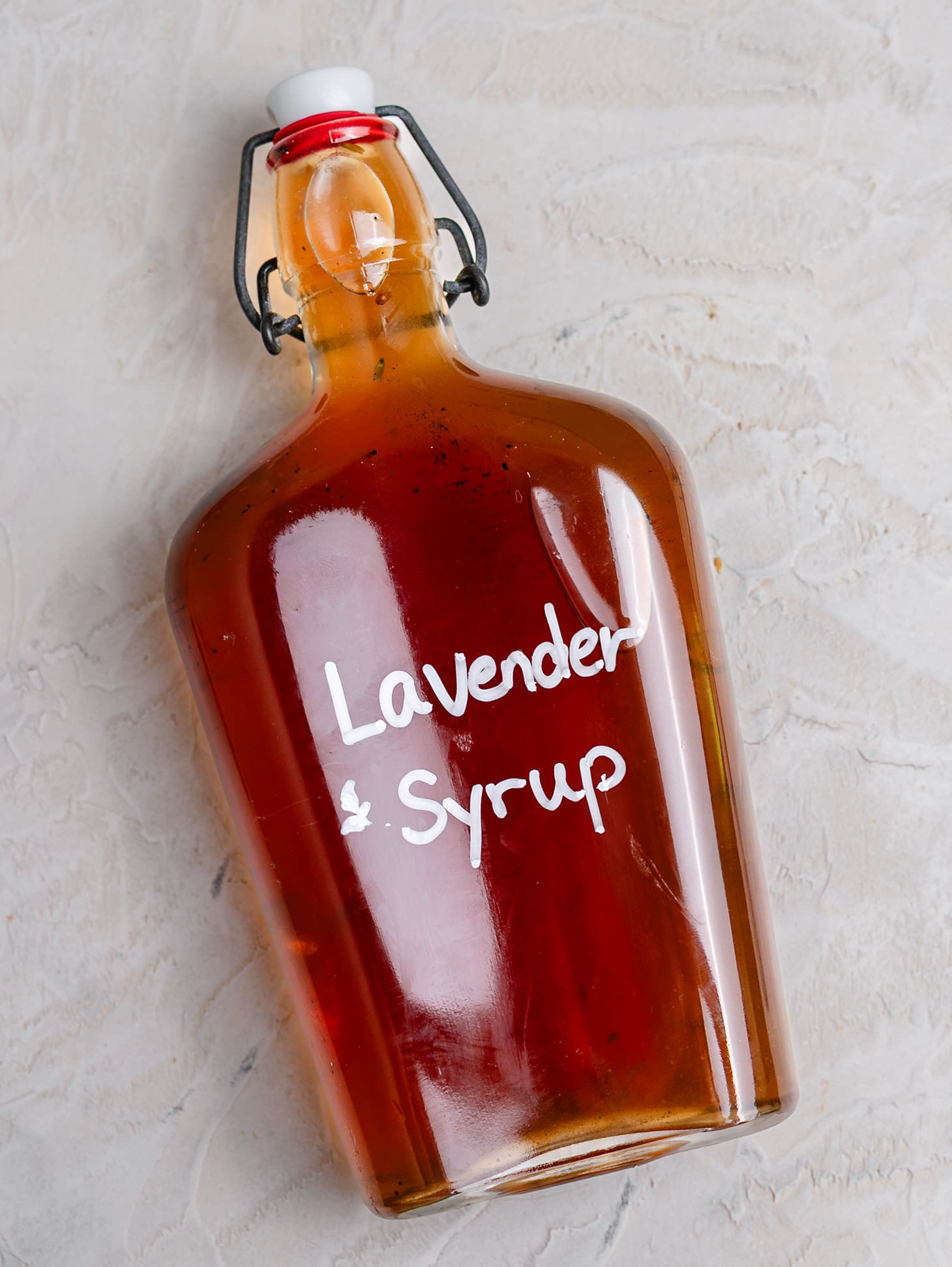 Glass bottle of homemade lavender syrup filled with golden amber syrup and labeled "Lavender Syrup" in white lettering. The bottle sits on a rustic white wooden board scattered with dried lavender buds, alongside a blue linen napkin and coffee mug in the background. A cozy kitchen scene highlighting an easy homemade coffee syrup recipe.