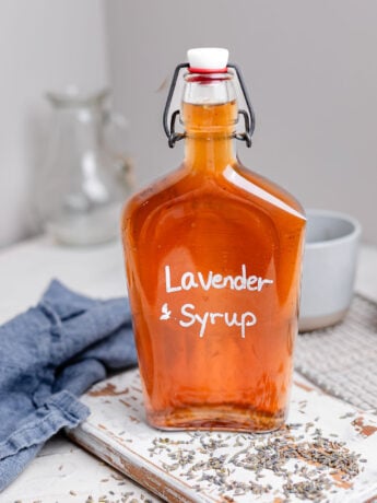 Glass bottle of homemade lavender syrup filled with golden amber syrup and labeled "Lavender Syrup" in white lettering. The bottle sits on a rustic white wooden board scattered with dried lavender buds, alongside a blue linen napkin and coffee mug in the background. A cozy kitchen scene highlighting an easy homemade coffee syrup recipe.